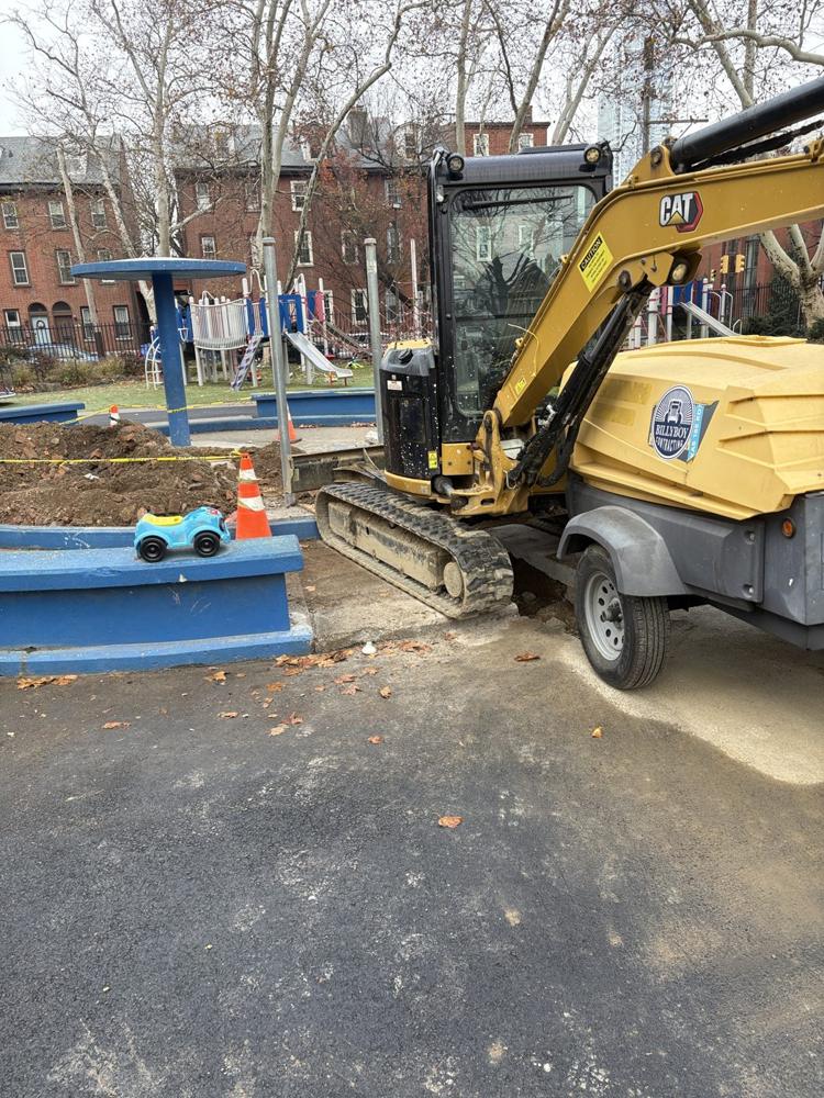 Excavator trenching the ground beside the spray ground and playground in Seger Park, December 2025, with a child's toy on the wall